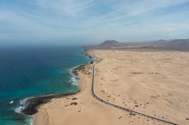İspanya, Fuerteventura 'daki Corralejo Doğal Parkı kum tepelerini geçerken çekilmiş yüksek çözünürlüklü hava fotoğrafı. Görüntü, altın kum tepeciklerinden oluşan engin bir çöl manzarasını kesen düz bir asfalt yolu gösteriyor.