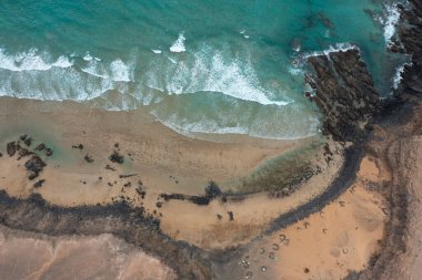 Kanarya Adaları 'ndan Fuerteventura' daki Esquinzo Sahili 'nin vahşi güzelliğini gösteren dramatik bir hava fotoğrafı. Bulutlu gökyüzü, turkuaz suların ve altın kumların üzerine yumuşak, karamsar bir ışık salıyor..
