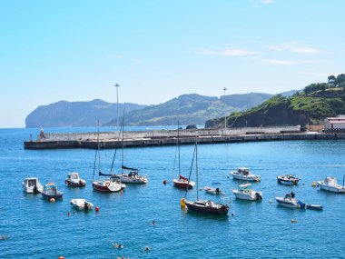 Tekneler ve yelkenli gemiler denizde, arkasında dağlar var.