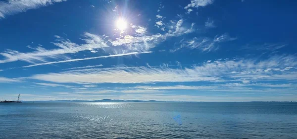 Mavi gökyüzünde parlak bir güneş ve denizin üzerinde uçan bulutlar. Gökyüzündeki uçakların deniz üzerindeki zıtlıkları.