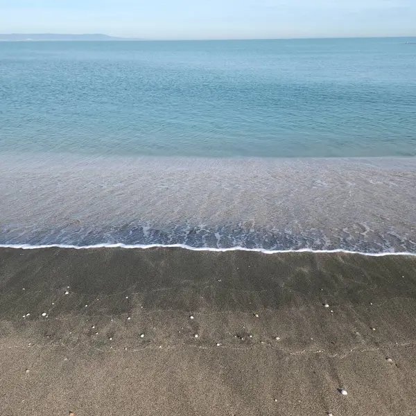 Deniz kıyısında bir gelgit dalgası vahşi bir kumsalın siyah kumu üzerinde yuvarlanıyor. Sakin denizin mavi suları