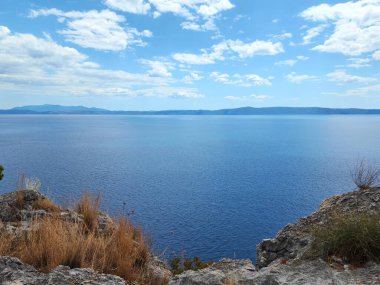Bulutlu gökyüzünün altında güneşli bir günde önplanda tepeler tarafından çerçevelenmiş parlak mavi denizin arka planı