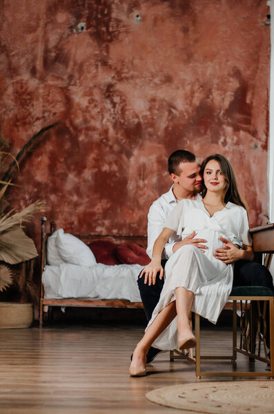 a pregnant girl and a man are sitting on a chair in an embrace in a brown room