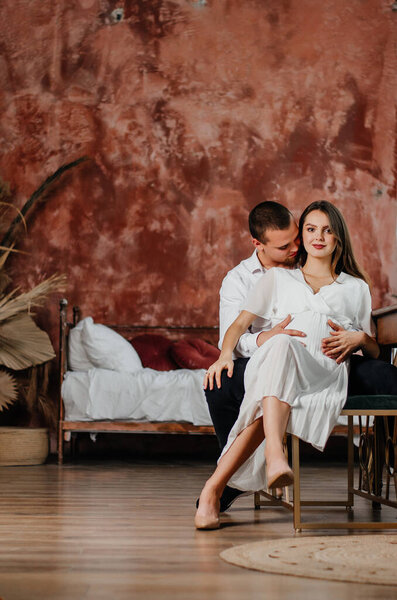 a pregnant girl and a man are sitting on a chair in an embrace in a brown room