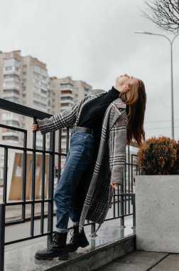 Gri paltolu ve kot pantolonlu bir kız korkuluklara tutunuyor.