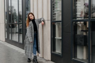 Bir kadın kaldırımda duruyor. Gri paltolu bir evin önünde.