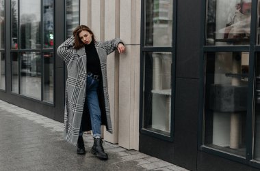 Bir kadın kaldırımda duruyor. Gri paltolu bir evin önünde.