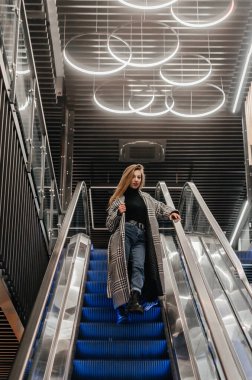 Paltolu bir kız yürüyen merdivenden aşağı iniyor.