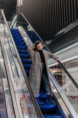 Paltolu bir kız yürüyen merdivenden aşağı iniyor.