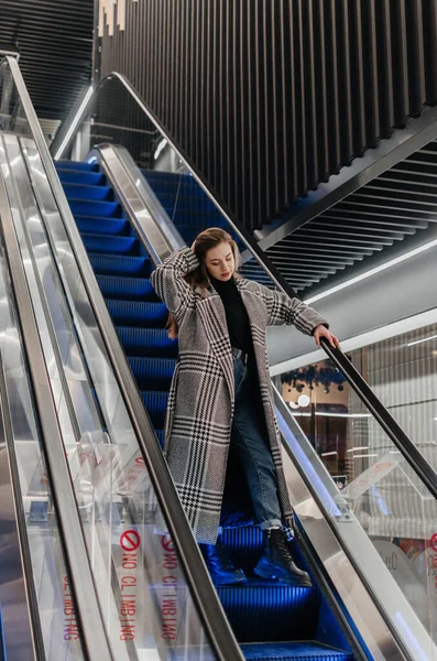 Paltolu bir kız yürüyen merdivenden aşağı iniyor.