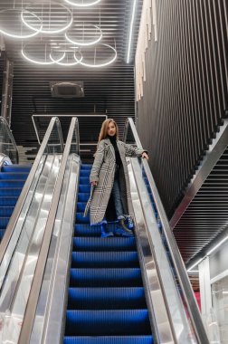 Paltolu bir kız yürüyen merdivenden aşağı iniyor.