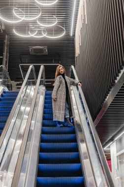 Paltolu bir kız yürüyen merdivenden aşağı iniyor.