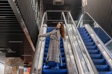 Paltolu bir kız yürüyen merdivenden aşağı iniyor.