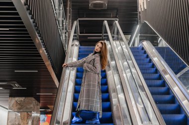 Paltolu bir kız yürüyen merdivenden aşağı iniyor.