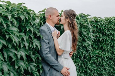 Gelin ve damat yeşil yapraklardan bir duvarın yanında kucaklaşıyor.