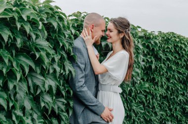 Gelin ve damat yeşil yapraklardan bir duvarın yanında kucaklaşıyor.
