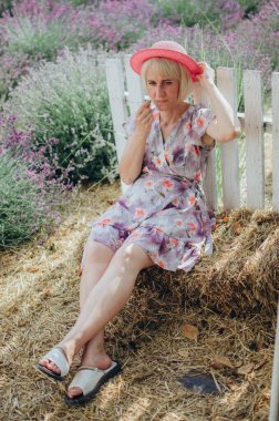 Lavanta tarlasında saman balyaları ve pembe hasır şapkalı, çiçekli elbiseli sarışın kadın.