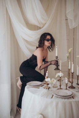 Siyah tulumlu zarif kadın elinde mumlar ve kumaşla klasik bir masada poz veriyor.