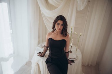 Siyah elbiseli kadın, masaya yaslanmış, karamsar ışıklandırmalı.