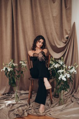 Zambak ve kumaşla çevrili bir kadın yerde oturuyor.