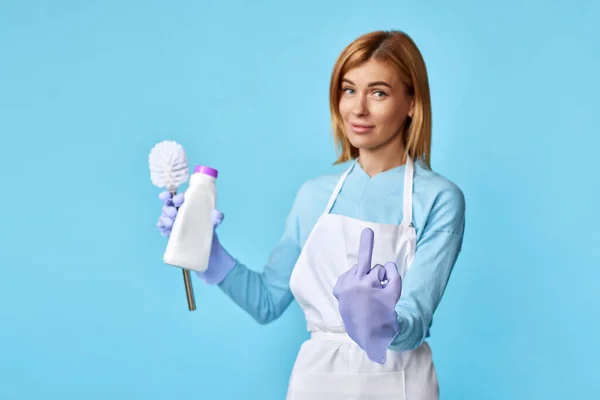 protest woman in rubber gloves and cleaner apron holding toilet brush and bottle of detergent and showing fuck you on blue background.