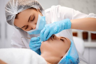 Güzellik kliniğinde mekanik yüz temizleme işlemi sırasında genç bir kadın. Kozmetik uzmanı siyah noktalardan ve sivilcelerden alınan çelik aletlerle derinin temizlenmesi için işlem yapıyor.