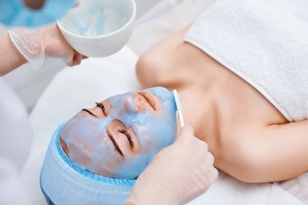 美容师在温泉沙龙的美女脸上涂上蓝色海藻酸钠面膜 面部皮肤光滑柔软 — 图库照片