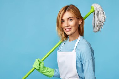 Yeşil lastik eldivenli güzel bir kadın ve temiz önlüklü mavi arka planda paspas tutuyor..