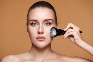 Güzel genç bir kadın bej arka planda profesyonel makyaj fırçasıyla pudra sürüyor.