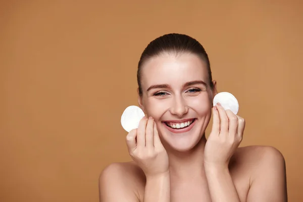 laughing young woman holding two white cotton disks pad with micellar water on beige background