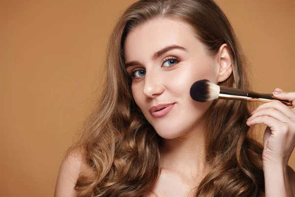 pretty curly woman applying blush on cheeks with make-up brush on beige background