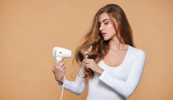 lovely woman with strong healthy curly hair using white hair dryer on beige background