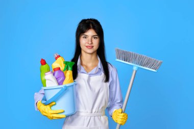Sarı lastik eldivenli, temiz önlüklü, elinde bir kova deterjan ve mavi arka planda süpürge tutan güzel bir kadın..