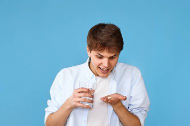 Genç yakışıklı adam mavi arka planda hap ve bir bardak su tutuyor. Sağlık, tedavi kavramı