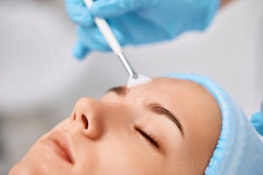 Güzellik uzmanı maskeyi güzellik salonundaki güzel genç bayanın yüzüne takıyor. Yaşlanma önleyici, yüz bakımı konsepti.