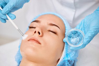 Güzellik uzmanı maskeyi güzellik salonundaki güzel genç bayanın yüzüne takıyor. Yaşlanma önleyici, yüz bakımı konsepti.
