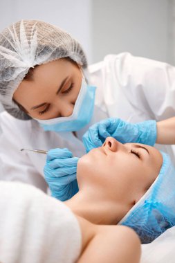 Güzellik kliniğinde mekanik yüz temizleme işlemi sırasında genç bir kadın. Kozmetik uzmanı siyah noktalardan ve sivilcelerden alınan çelik aletlerle derinin temizlenmesi için işlem yapıyor.