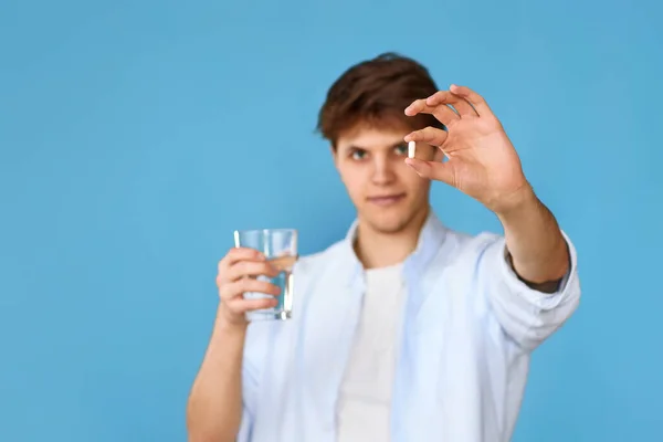 年轻英俊的男子拿着白色石碑和蓝色背景的杯子水 专注于药丸 — 图库照片