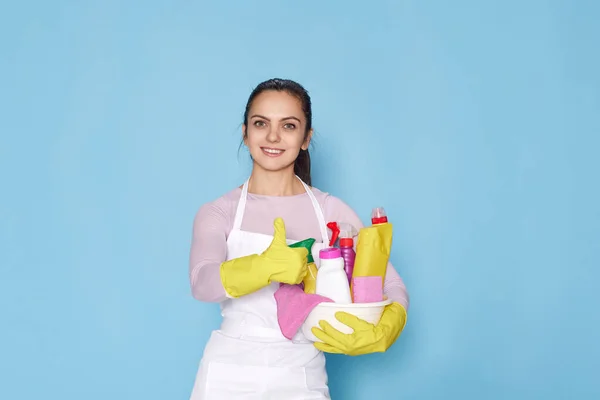 happy caucasian woman in gloves and cleaner apron holding bucket of detergents and showing Ok gesture on blue background.