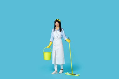 Profesyonel temizlikçi kadın lastik eldivenler ve temiz önlükle mavi arka planda paspas ve kova tutuyor. Tam uzunluk