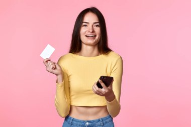 Pembe arka planda izole edilmiş cep telefonu ve plastik kredi kartı kullanan sarı elbiseli heyecanlı bir kadın. İnternetten alışveriş.