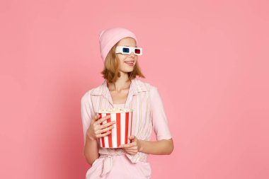 Pembe tişörtlü, şapkalı, elinde patlamış mısırla pembe arka planda duran güzel sarışın kız. Boşluğu kopyala