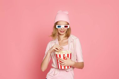 3 boyutlu gözlüklü çekici sarışın kadın pembe arka planda izole edilmiş patlamış mısır kovası tutuyor.