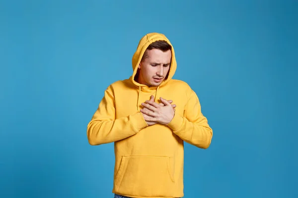 young man in yellow sweatshirt with hands on chest with closed eyes isolated on blue background.