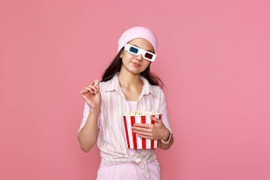 Pembe şapkalı gülümseyen beyaz kız film izliyor, elinde pastel pembe arka planda izole edilmiş patlamış mısır kovası var..