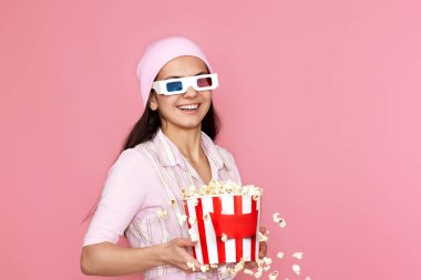 Üç boyutlu gözlüklü neşeli kadın, elinde patlamış mısır kovası olan şapkalı ve pastel pembe arka planda izole bir şekilde atlayan..