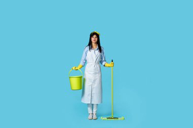 Lastik eldivenli güzel bir kadın ve temiz önlüklü mavi arka planda paspas ve kova tutan. Tam uzunluk