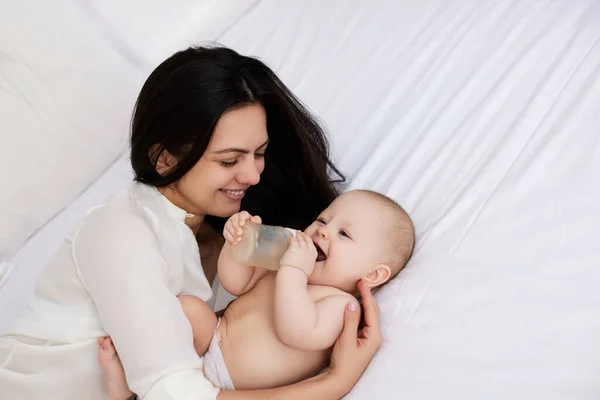 happy mother feeding her newborn baby in a white bedroom. mom holding a bottle of formula milk
