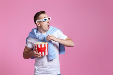 Üç boyutlu gözlüklü, korkmuş yakışıklı adam pembe arka planda patlamış mısır kovası tutuyor.