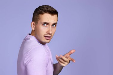indignant man in t-shirt asks a question and looking back over his shoulder on purple background. What do you want. copy space clipart